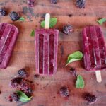 Berry popsicles on a wood board surrounded by blackberries and mint leaves.