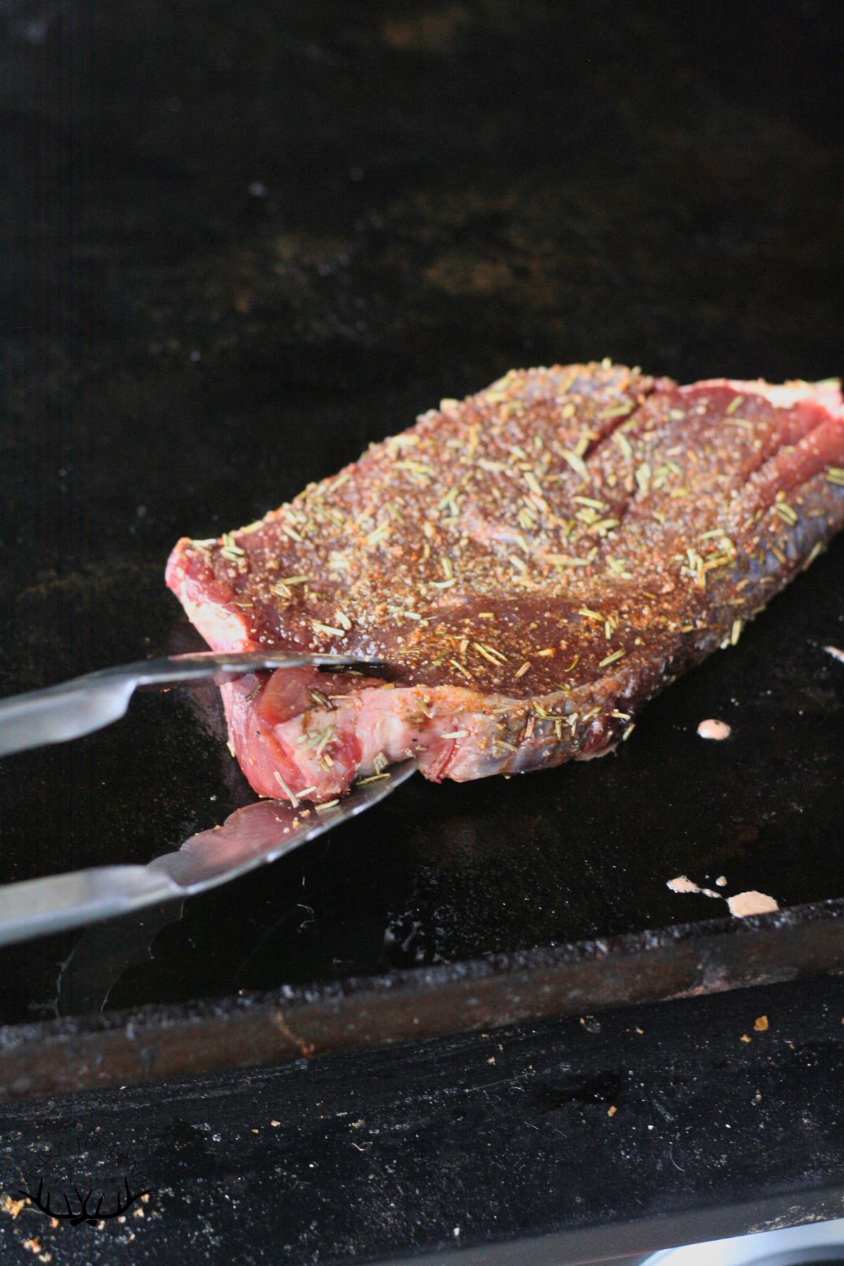 Tongs flipping a moose steak on a Blackstone griddle.